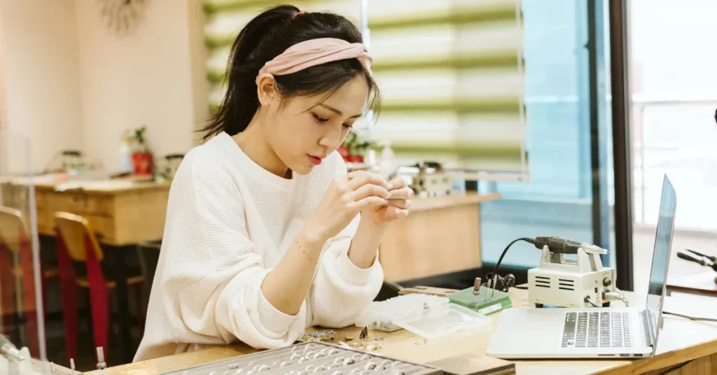Woman crafting DIY acrylic gem jewelry at a worktable with tools, gemstones, and a laptop.