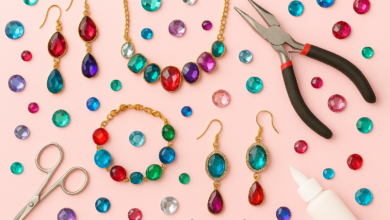 A flat lay of handmade DIY acrylic gem jewelry including necklaces, earrings, and bracelets with colorful flat-back gems, tools, and glue on a pastel background.
